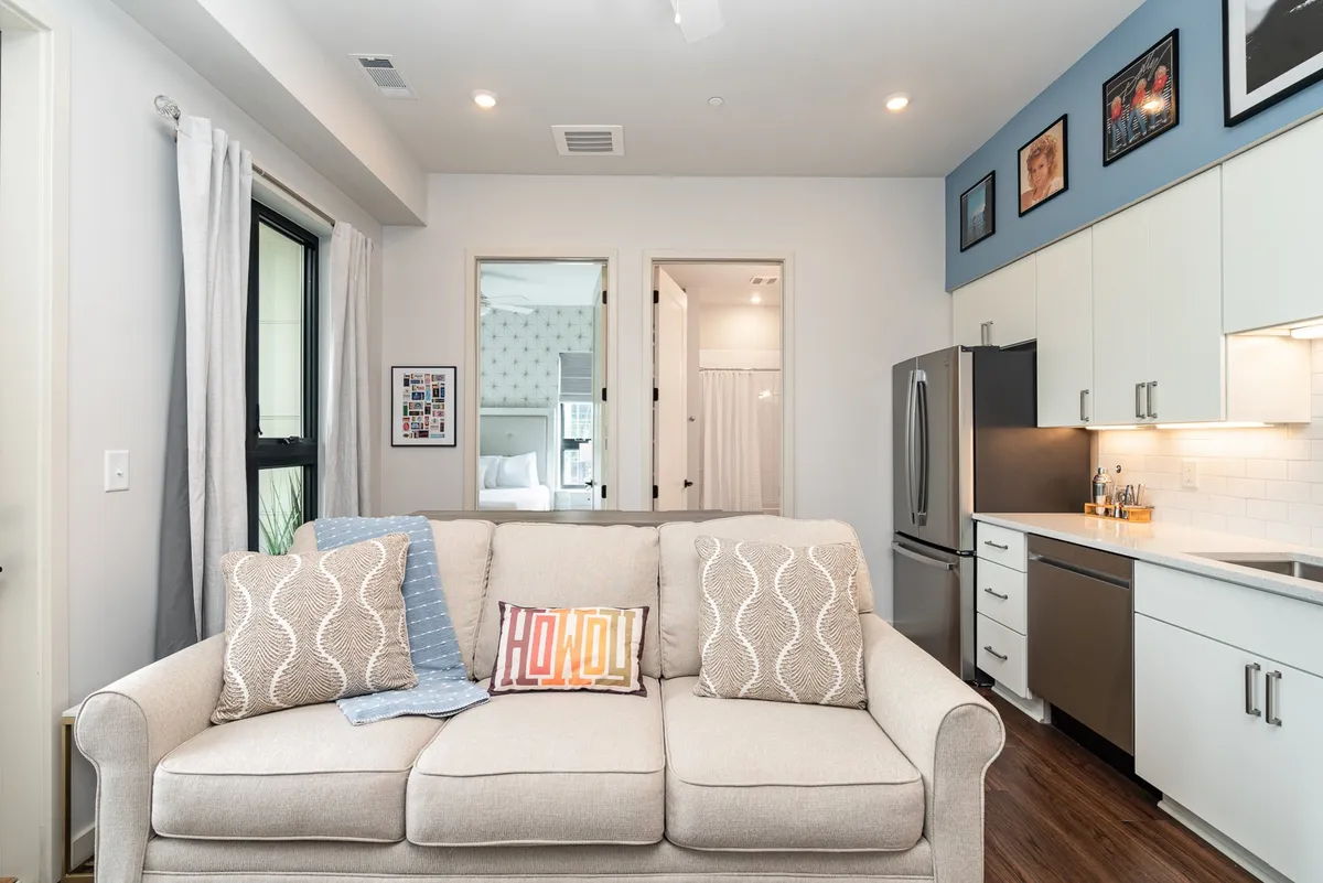 This image showcases a bright, open-concept living space with a plush sofa, kitchen area, decorative artwork, and doorways leading to adjacent rooms. The layout emphasizes the flow between the lounge and kitchen, with an inviting ambiance perfect for relaxing or entertaining guests.