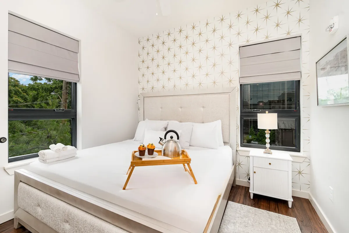 A beautifully styled bedroom with a plush bed, breakfast tray, large windows, decorative wallpaper, and a softly glowing lamp. Natural light floods the room, creating a serene and cozy ambiance perfect for relaxation.