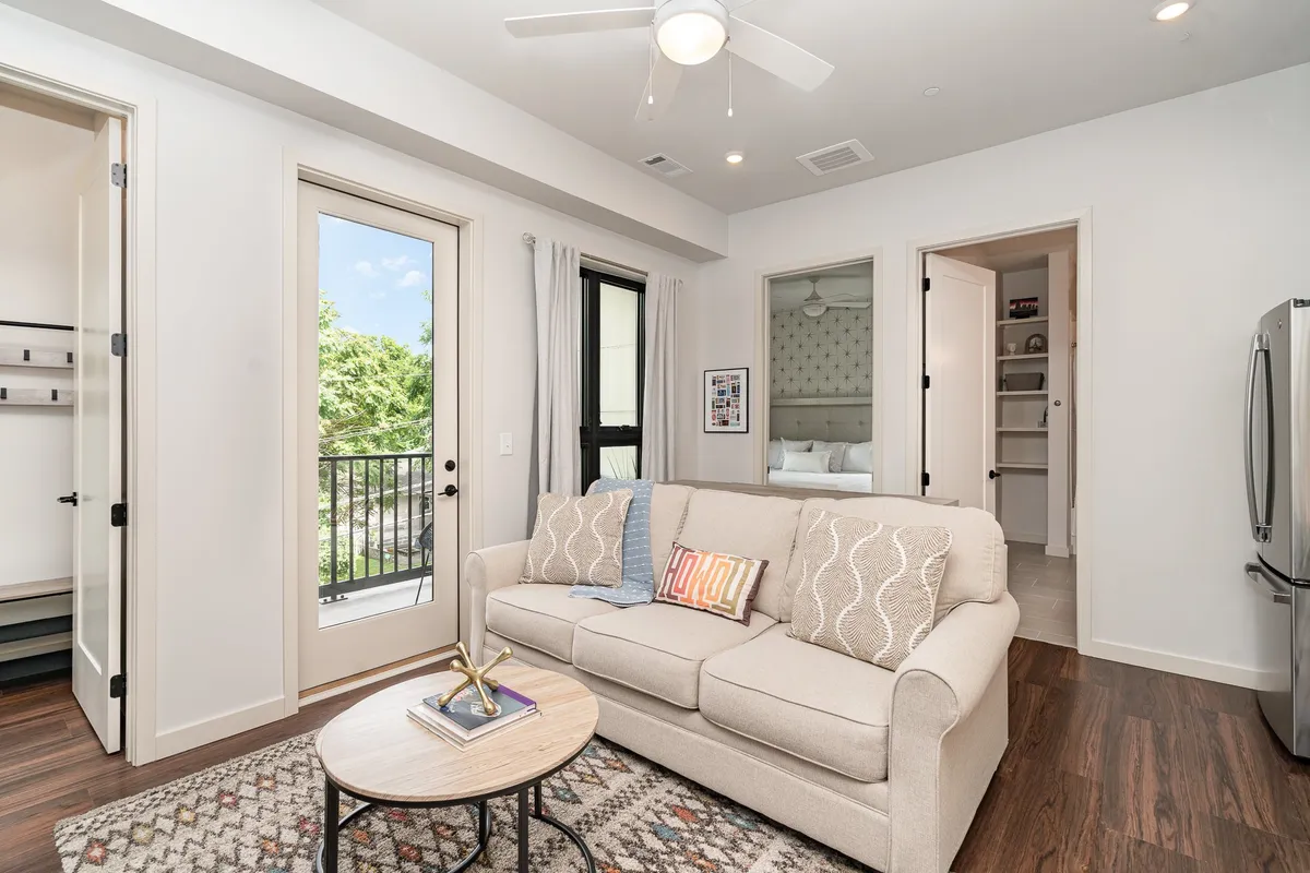A cozy seating area with a plush sofa and coffee table, framed by large balcony doors that flood the space with natural light. The artwork on the wall and sunrise view from the balcony add warmth, while the layout invites conversation and relaxation.
