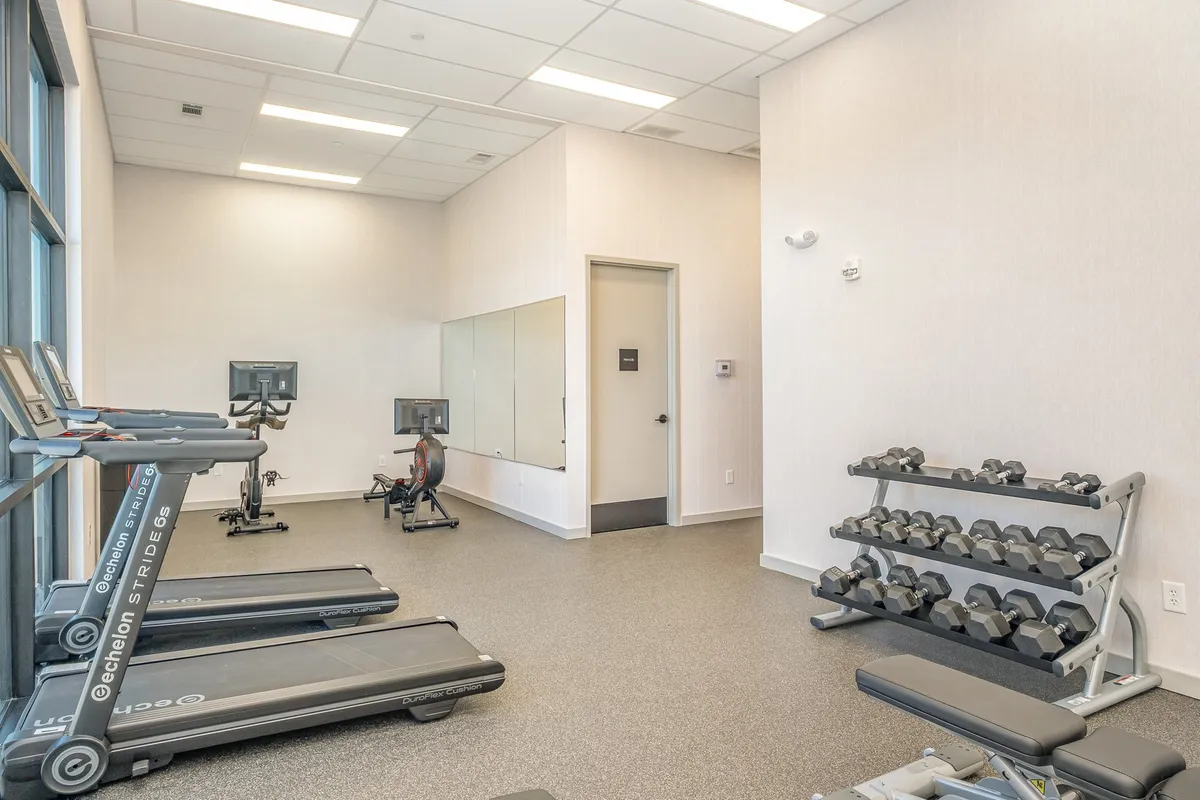 A bright, modern gym featuring treadmills, a dumbbell rack, an exercise bike, and mirrors. The space is designed for both individual workouts and group fitness, emphasizing a clean and motivating environment.